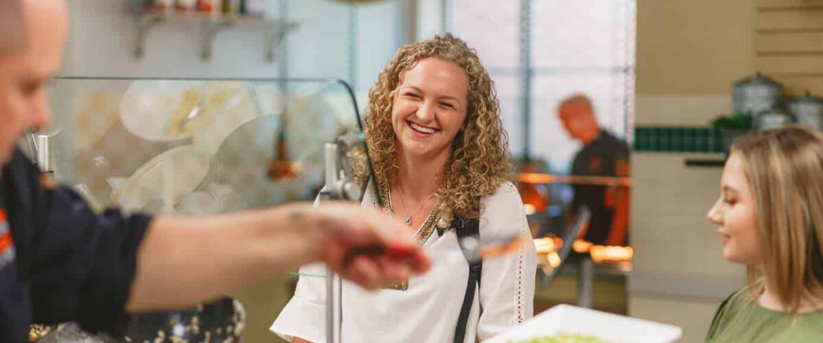 Student smiling while waiting for dish to be prepared in cafeteria line